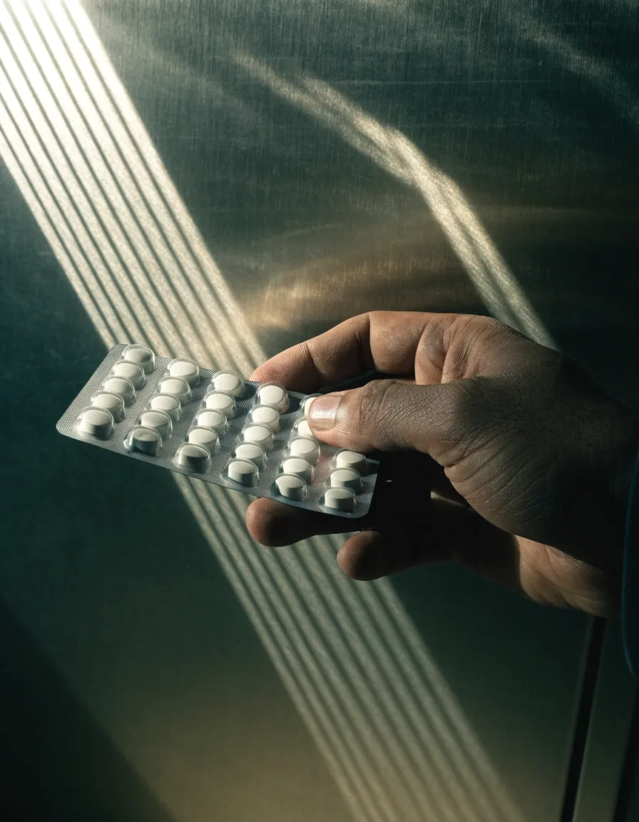 A hand comparing a blister pack of capsules against a plain panel under hard slat light over a dark counter