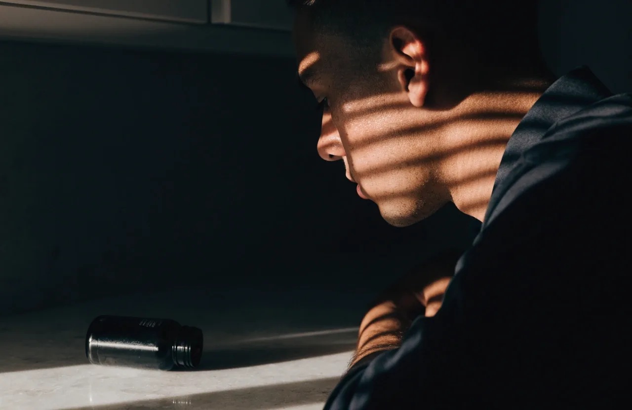 A man in profile at a kitchen counter in hard slatted morning light with a matte unmarked bottle in front of him